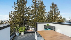 Una terraza en la azotea con una mesa de madera, sillas blancas y plantas en macetas.
