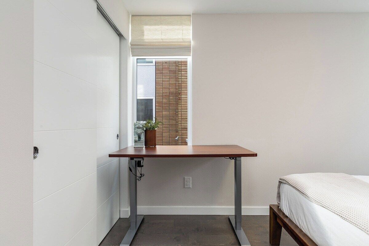 A small room with a bed, a wooden desk, and a window with blinds.