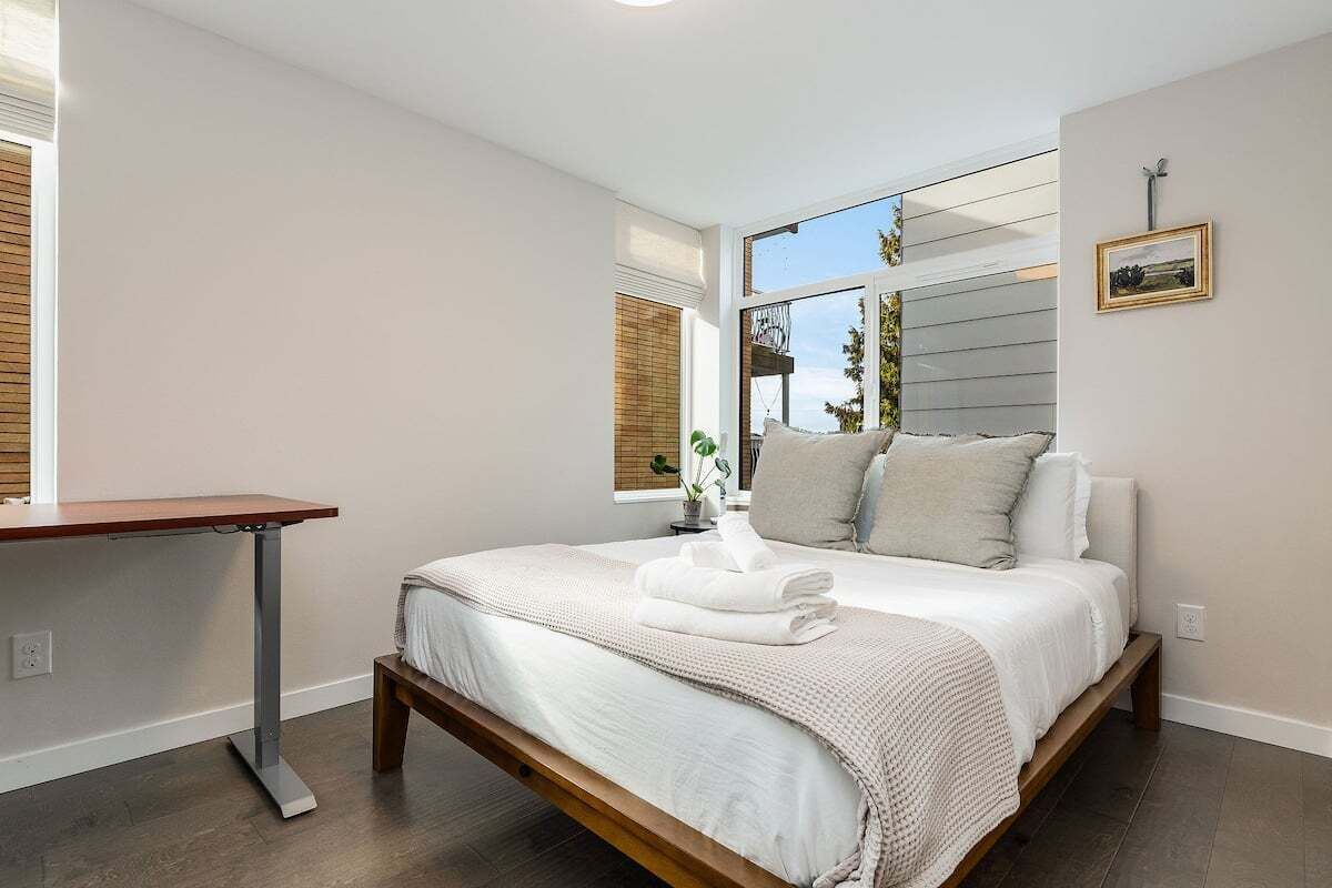 Un dormitorio con cama, mesita de noche, ventana y un cuadro en la pared.