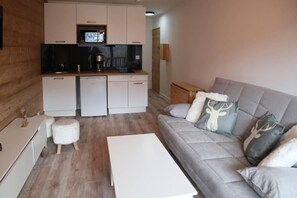 A modern living room with a grey sofa, white coffee table, and a kitchen area with white cabinets and appliances.
