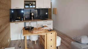 Une cuisine compacte équipée d’un four à micro-ondes, d’une machine à café et d’un petit réfrigérateur. On y trouve une table en bois avec une chaise et un coussin blanc et moelleux.
