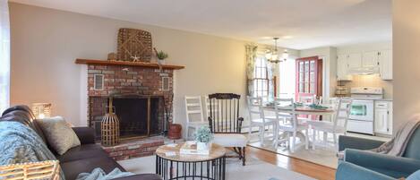 A living room with a brick fireplace, a dining area with white chairs, and a kitchen with white cabinets.