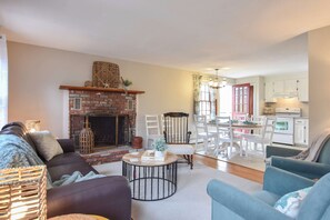 A living room with a brick fireplace, a dining area with white chairs, and a kitchen with white cabinets. - Minutes to Oceanside Beaches! (Eastham)