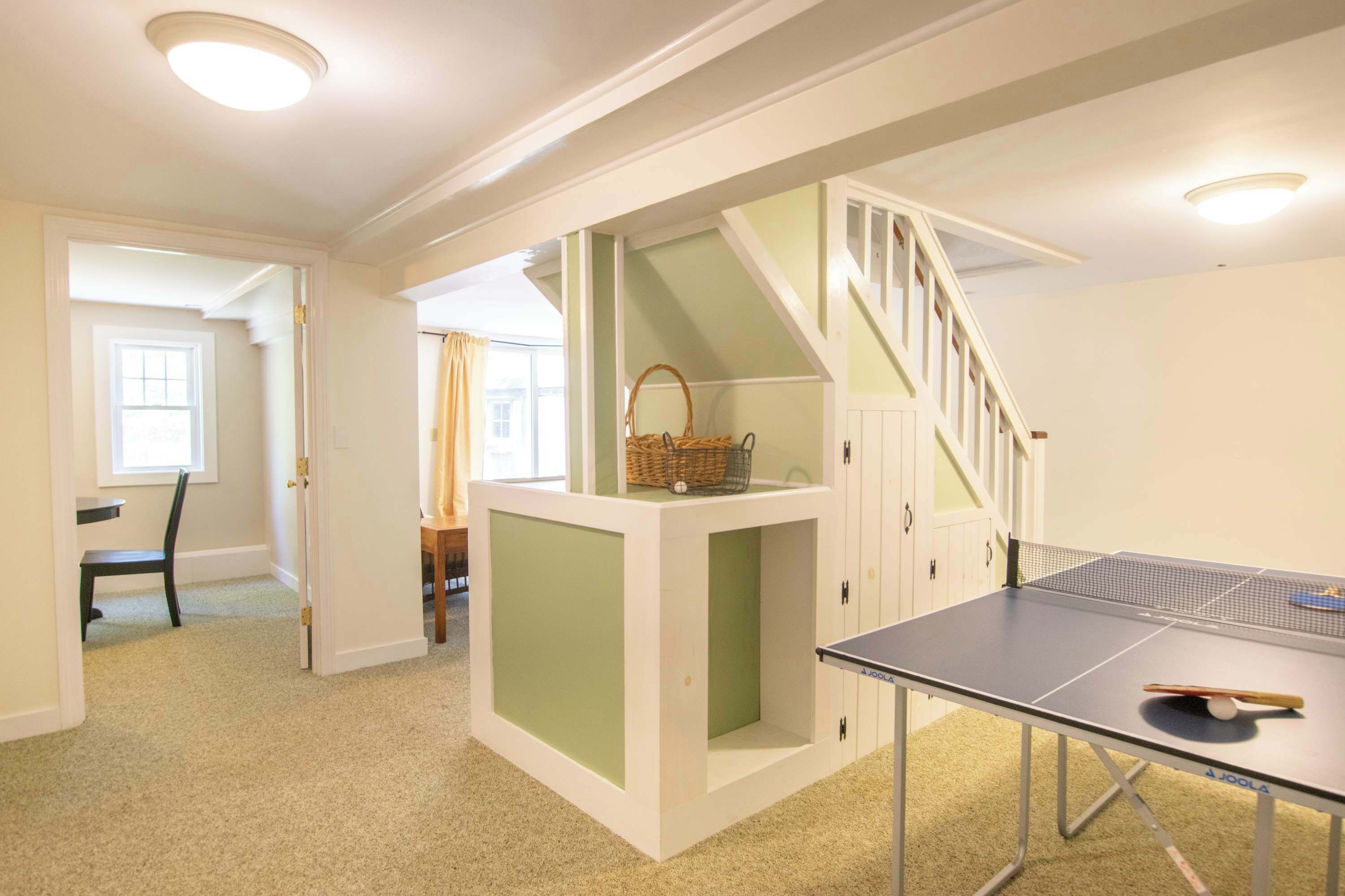 Une pièce avec une table de ping-pong, une chaise, une petite table avec un panier et un escalier.