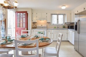 A modern kitchen with white cabinets, a stainless steel refrigerator, and a dining area with a wooden table and white chairs.