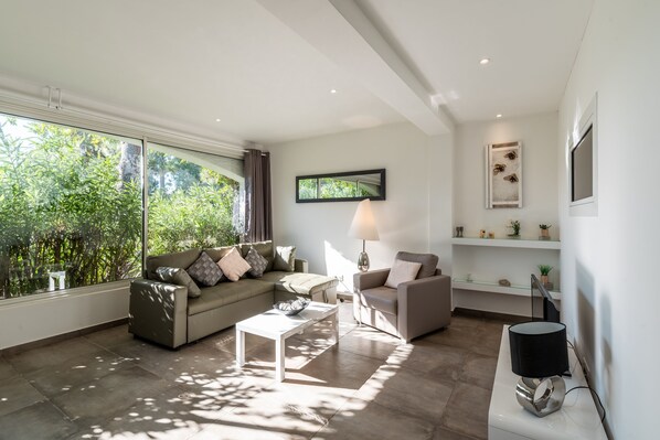 A modern living room with a sofa, armchair, and coffee table, large windows with a view of greenery, and a wall-mounted TV.