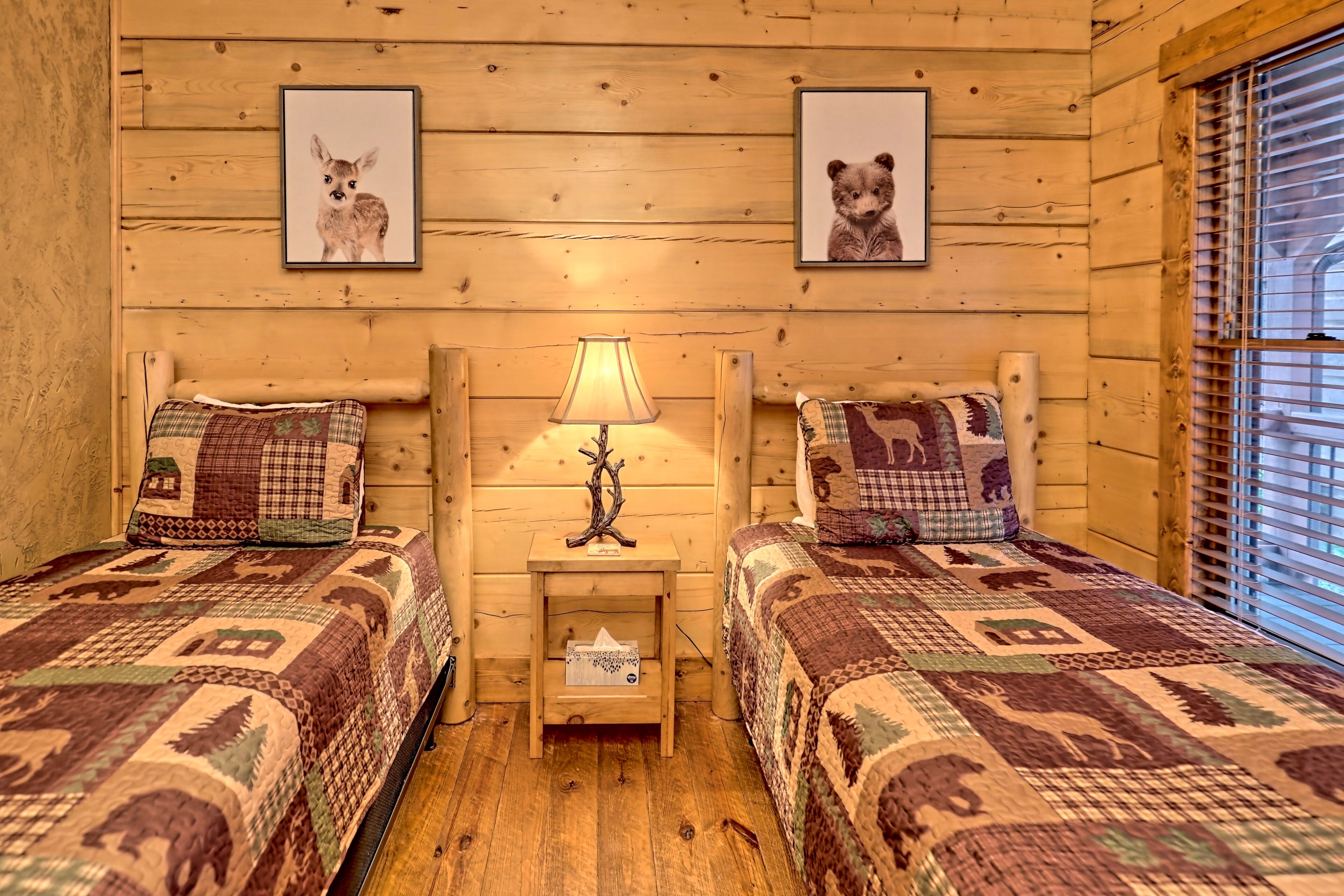 A cozy cabin room with two beds, each with a patterned quilt, a small bedside table with a lamp, and framed pictures of animals on the wall.