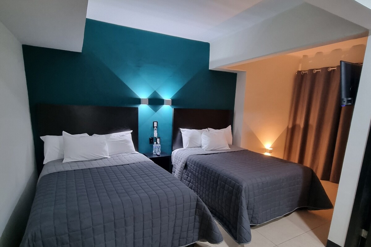 A hotel room with two beds, a nightstand with a lamp, and a bathroom visible through an open door.