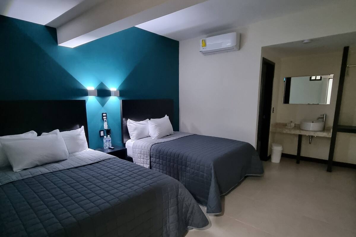 A hotel room with two beds, a bathroom with a sink and mirror, and an air conditioning unit on the wall.