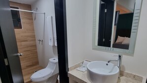 Un baño moderno con inodoro blanco, lavamanos con espejo y zona de ducha visible a través de una puerta abierta.