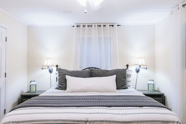 A neatly made bed with a striped bedspread, two bedside lamps, and a window with sheer curtains. - Luxurious cabin with fire pit, kayaks, and dock. (Arley)