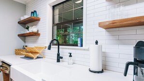 A kitchen with white countertops, wooden shelves, a window, and a coffee maker. - Luxurious cabin with fire pit, kayaks, and dock. (Arley)