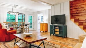 A spacious living room with a wooden floor, a dining area with a table and chairs, a television on a stand, and a staircase with wooden railings. - Luxurious cabin with fire pit, kayaks, and dock. (Arley)