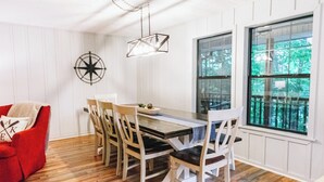 A dining area with a wooden table and chairs, a red armchair, and a large window with a view of trees. - Luxurious cabin with fire pit, kayaks, and dock. (Arley)