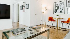 Una sala de estar moderna con un televisor de pantalla plana, una mesa de centro de vidrio con libros y un control remoto, dos sillas naranjas alrededor de una mesita y cuadros en la pared.