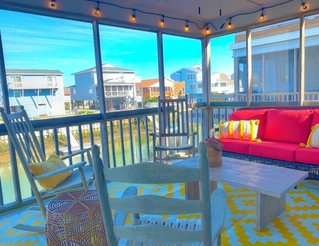 A sunlit balcony with wicker furniture, a red sofa, and a view of neighboring houses.