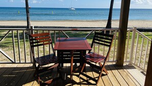 Terraza de madera con dos sillas y una mesa, con vista a una playa y una vista despejada del océano, con un velero a lo lejos.