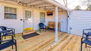Een overdekt terras met houten vloer, twee zwarte wicker stoelen, een houten bank en een aan de muur bevestigde televisie.