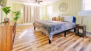 Een slaapkamer met een bed, houten vloer, een bureau, een stoel, een plant, een plafondventilator en een raam met gordijnen.