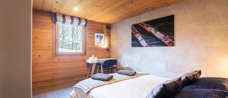 A wooden cabin bedroom with a bed, a desk, and a window with a view of trees.