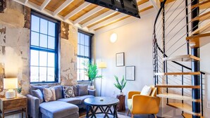 Un salón acogedor con sofá, sillón y una mesita redonda. Se observa una escalera con barandilla de metal y una planta en la esquina.