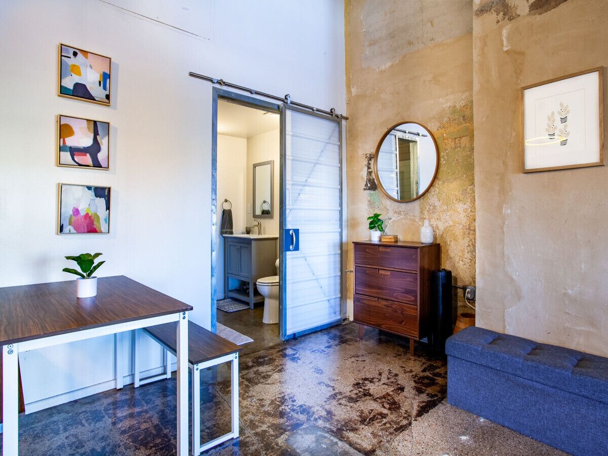 A room with a sliding door leading to a bathroom, a small table with a plant, a dresser, and a round mirror on the wall.