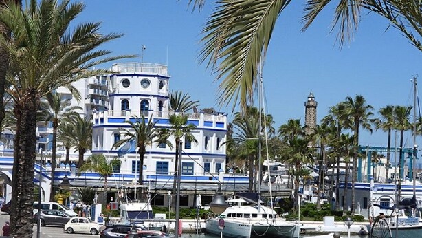 Un bâtiment situé sur la côte, doté d’une tour horloge, et entouré de palmiers et de bateaux.