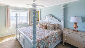 A bedroom with a four-poster bed, a ceiling fan, a nightstand with a lamp, and a view of the ocean through a window. - Recently renovated condo in Cherry Grove area of N. Myrtle Beach (North Myrtle Beach)