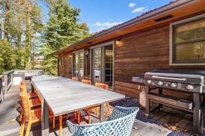A wooden cabin with a deck, a long table, orange and blue chairs, and a Blackstone grill.