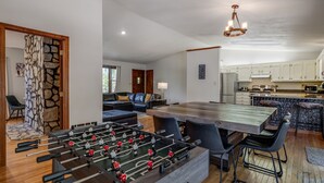 A modern living room with a foosball table, a dining area with a large wooden table, and a kitchen with white cabinets.