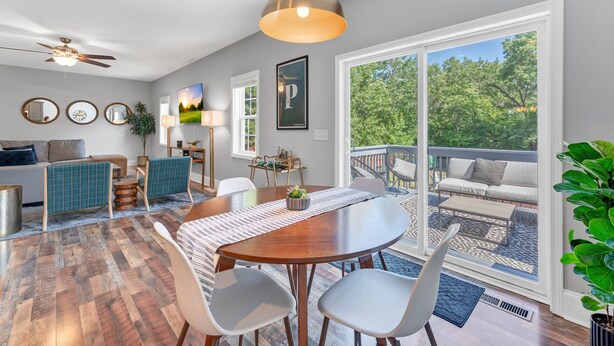 A spacious living room with a large wooden dining table, white chairs, and a ceiling fan. There is a balcony with outdoor seating and a view of greenery.