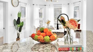 Eine Küche mit Marmorarbeitsplatte, Obstkorb, Blumenvase und einem Essbereich im Hintergrund.