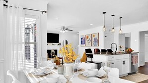 Ein moderner Essbereich mit einem weißen Tisch, gedeckt für eine Mahlzeit, einer Küche im Hintergrund und Blick ins Freie durch ein Fenster mit weißen Vorhängen.