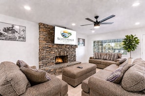 A living room with a stone fireplace, a ceiling fan, and a sign for Scottsdale Stays LLC. - Scottsdalestays Shangri-La 10BR-5BA on 1/2 Acre-Resort Pool w/Cabana-Pickleball (Phoenix)