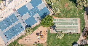 Ein Luftbild einer Sportanlage mit mehreren Plätzen, darunter Tennis und Ruckelbahnen, umgeben von Grünflächen und Palmen.