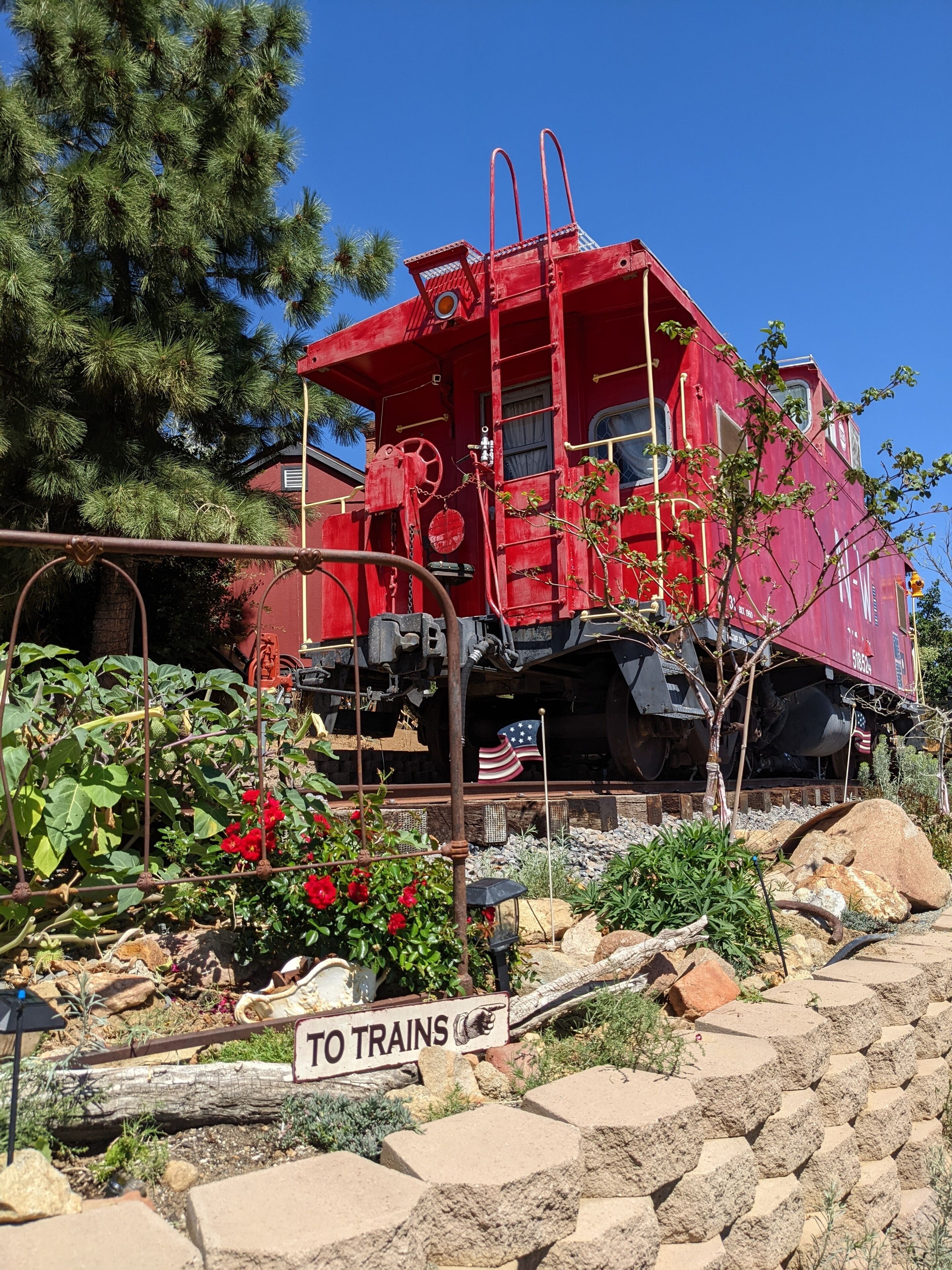 Una locomotora estacionada con exterior rojo, rodeada por un jardín con diversas plantas y un letrero que dice 'TO TRAINS'.