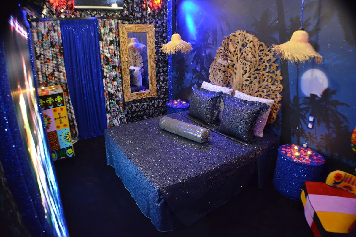 A room with a bed covered in a glittery blue bedspread, a mirror, a decorative headboard, and a small table with candles.