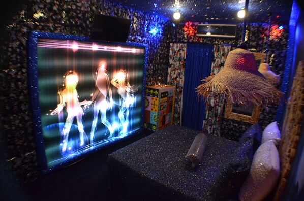 A room with a large screen displaying a performance, a bed with a decorative headboard, and a small table with a box and a straw hat.