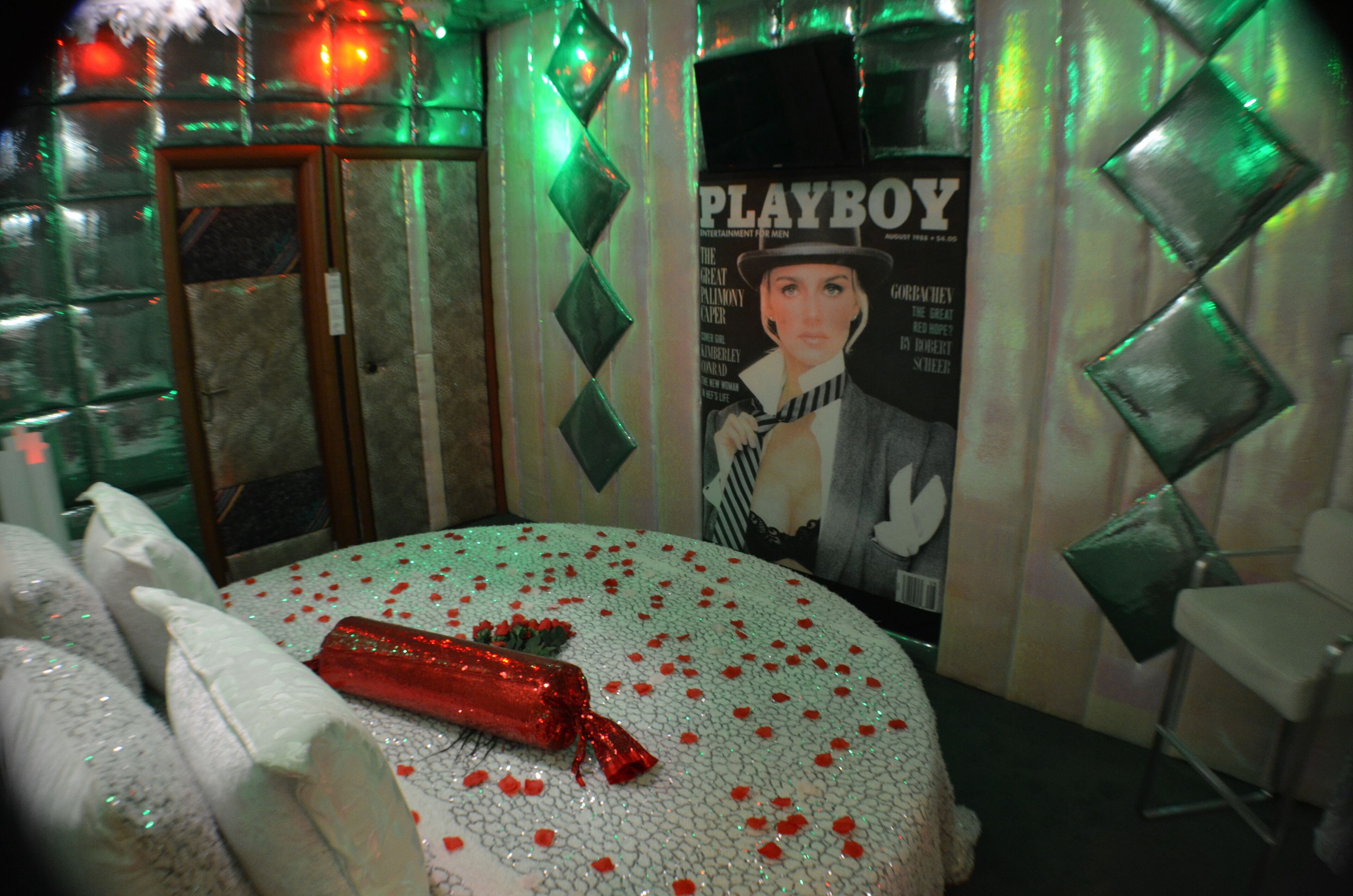 A room with a round table covered in rose petals and a red gift. There is a poster on the wall and a chair to the side.