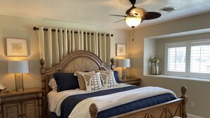 A bedroom with a large bed, wooden headboard, two bedside tables with lamps, a bench, a ceiling fan, and a window with blinds. - Beachside Daytona Beach Shores 5 min walk to beach, restaurants and more! (Port Orange)