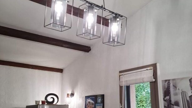 A kitchen with a modern chandelier, a refrigerator, a dining table with a placemat, and a window with a view of greenery. - Comfortable air-conditioned studio in a quiet villa 15 km from Cannes (Peymeinade)