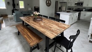 A spacious dining area with a wooden table and benches, a high ceiling with a hanging light fixture, and a kitchen in the background. - Coastal Cape Hideaway - Outdoor Shower & Fire-pit! (Mashpee)