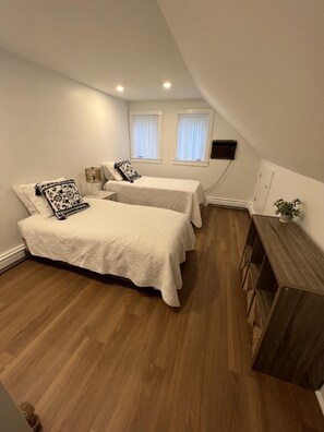 A small, modern bedroom with two beds, a wooden floor, a bedside table, and a small plant. - Coastal Cape Hideaway - Outdoor Shower & Fire-pit! (Mashpee)