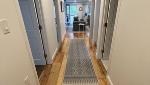 A hallway with wooden flooring, a patterned rug, and doors leading to a living area. - Coastal Cape Hideaway - Outdoor Shower & Fire-pit! (Mashpee)