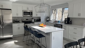 A modern kitchen with white cabinets, a stainless steel refrigerator, a microwave, a stove, and a coffee maker. There are bar stools and a fruit bowl on the island. - Coastal Cape Hideaway - Outdoor Shower & Fire-pit! (Mashpee)