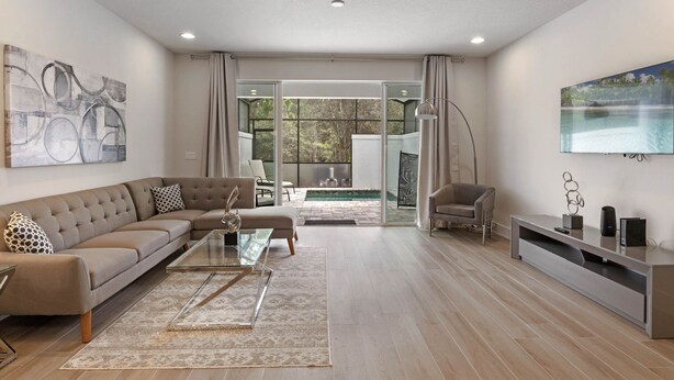Un moderno salón con sofá, mesa de centro y mueble para televisión. Hay una puerta de cristal que da a un balcón con piscina.