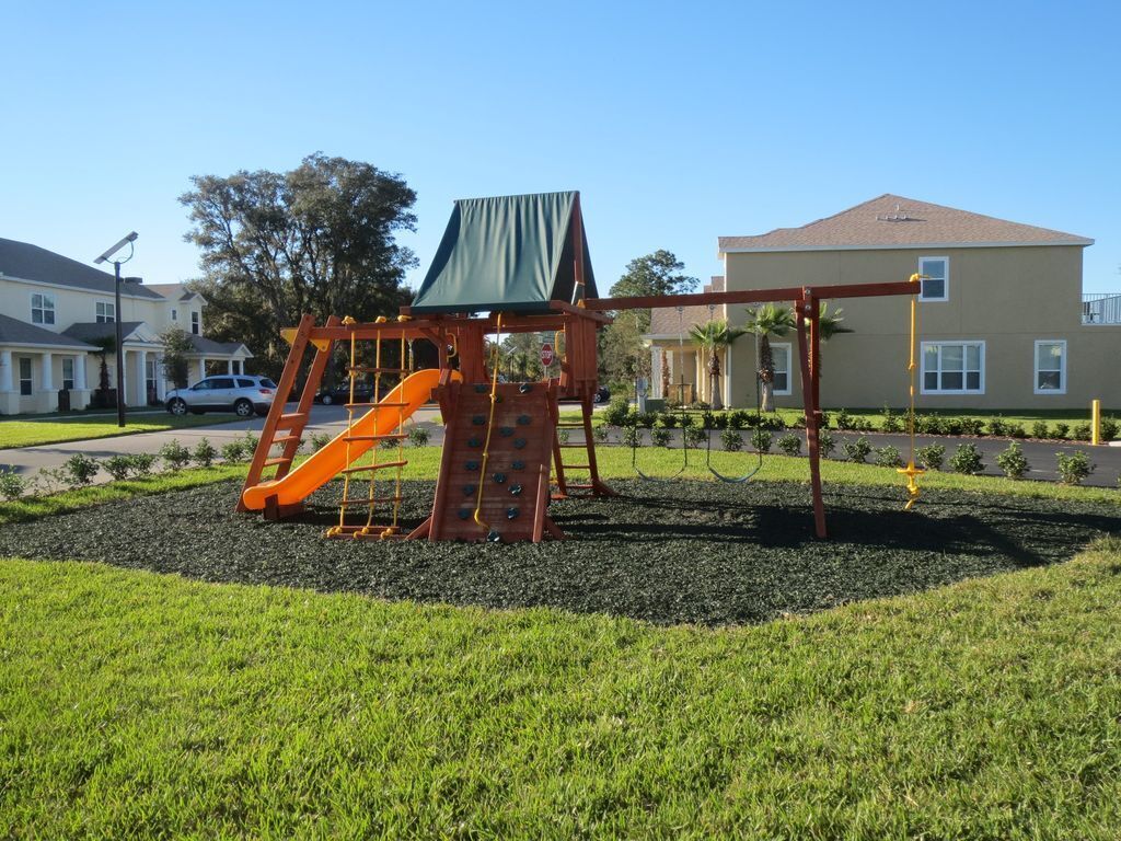 Un parque infantil con un tobogán y columpios, rodeado de césped y un edificio al fondo.