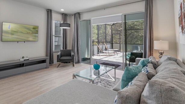 A modern living room with a grey sofa, a glass coffee table, and a flat-screen TV mounted on the wall. - Jose's Tikki Room near Disney (Clermont)