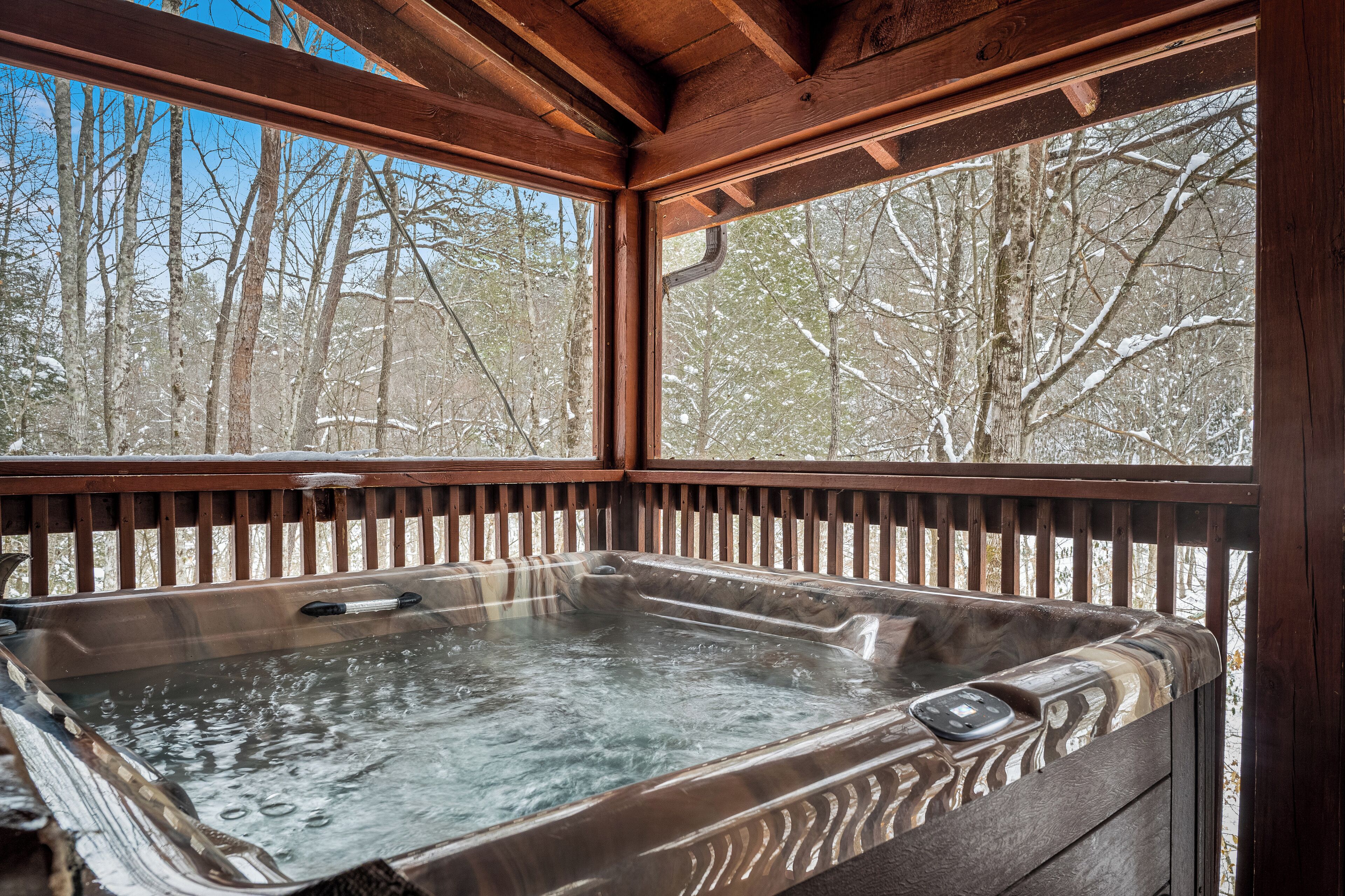 A hot tub with a wooden deck and windows overlooking a snowy forest.
