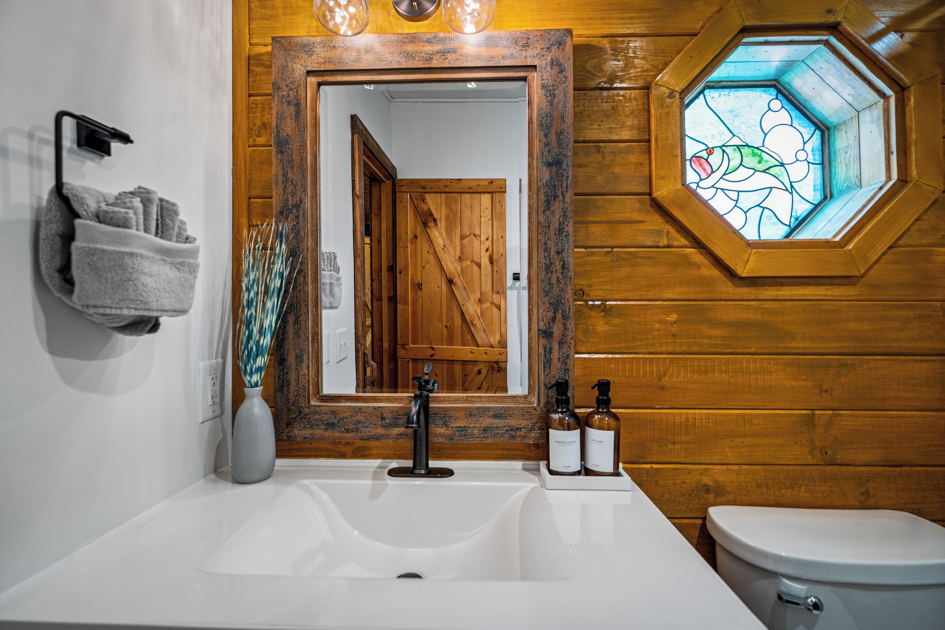 A bathroom with a wooden wall, a white sink, a mirror, a towel rack, a toilet, and a stained glass window.